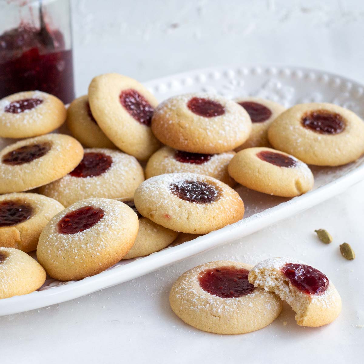 Cardamom Jam Thumbprint cookies in a white platter. In front of the platter are two jam cookies, and one has a bite in it. There are also two cardamom pods next to the cookies. On the back is a jar of raspberry jam.