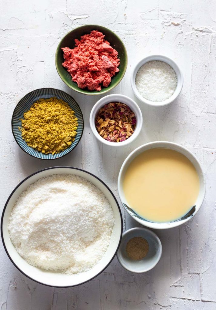 Ingredients for instant coconut ladoos (or cardamom coconut fudge balls). Ingredients clockwise from top are freeze dried strawberries, coconut, rose petals, condensed milk, cardamom powder, desiccated coconut and pistachio powder.