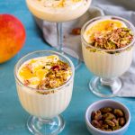 Three glasses of mango mousse on a blue background. The mango mousse cups are garnished with mango puree, pistachios and rose petals. On the top right corner is a mango, and on the bottom right corner is a small bowl of pistachios.