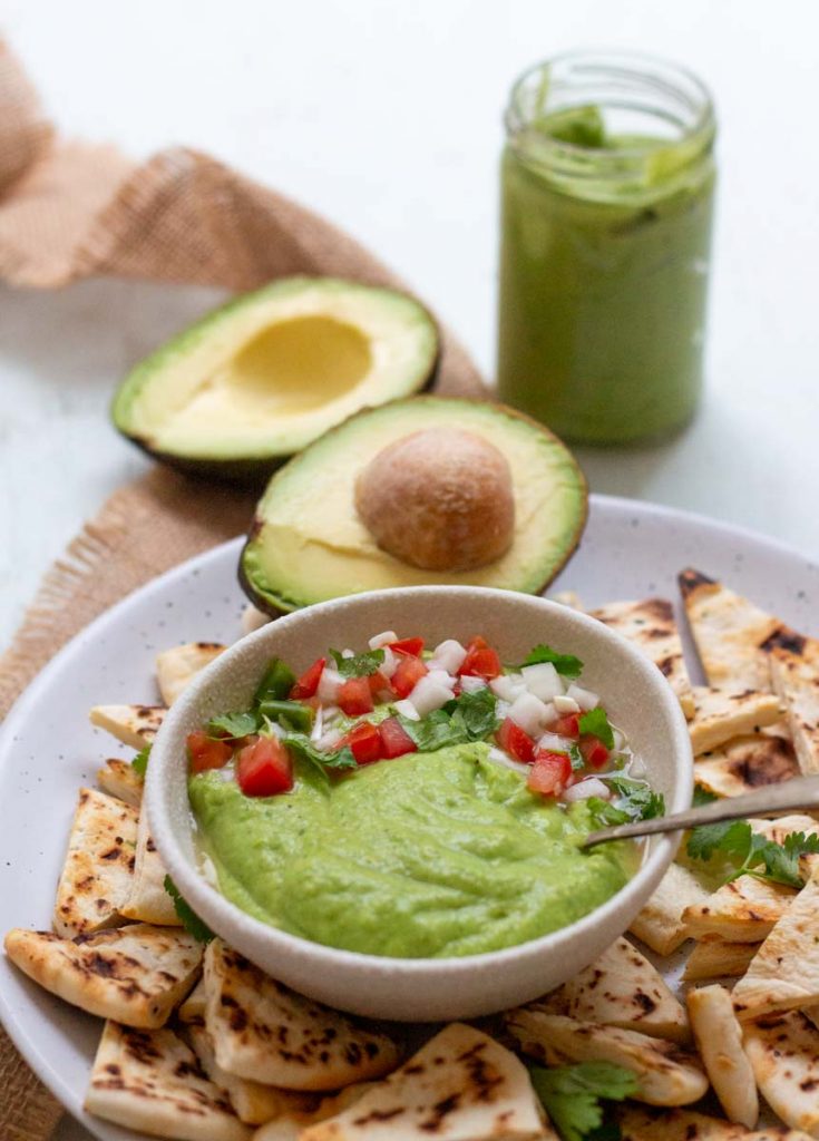 A plate of toasted pita chips. In the middle of the plate is a bowl of avocado green chutney with a spoon in it, and garnished with chopped coriander, tomatoes and onion. On the back of the plate is 1 avocado cut in half, and a jar of avocado green chutney in the background.