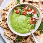 A plate of toasted pita chips. In the middle of the plate is a bowl of avocado green chutney with a spoon in it, and garnished with chopped coriander, tomatoes and onion.