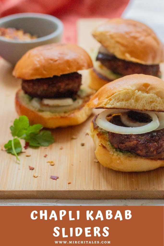 Three chapli kabab sliders on a wooden board with a rust colored napkin in the background and a small bowl of red chili flakes.