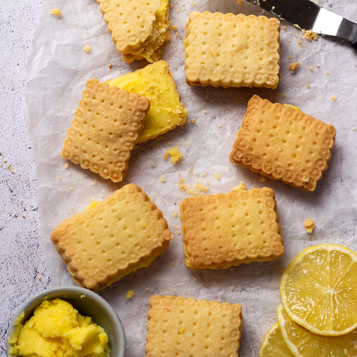 Lemon sandwich cookies laid out on parchment paper. On the bottom right are three lemon slices, and on the bottom left is a small bowl with lemon buttercream filling.