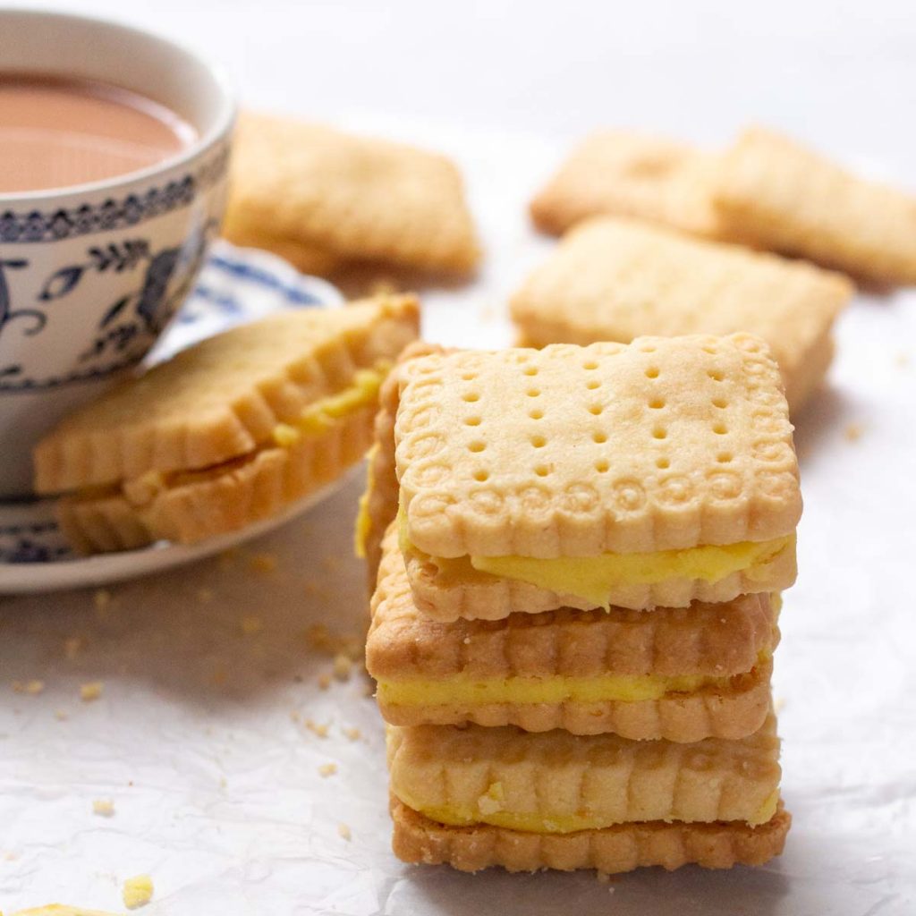 A stack of three lemon sandwich cookies with lemon buttercream filling. On the back in the top left corner is a cup of tea with a saucer on which a biscuit is kept. In the background are a few lemon sandwich cookies as well.